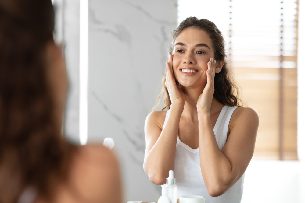 Young woman looking in the mirror smiling with hands on face, happy with her Restylane Lyft treatment Young woman looking in the mirror smiling with hands on face, happy with her Restylane Lyft treatment