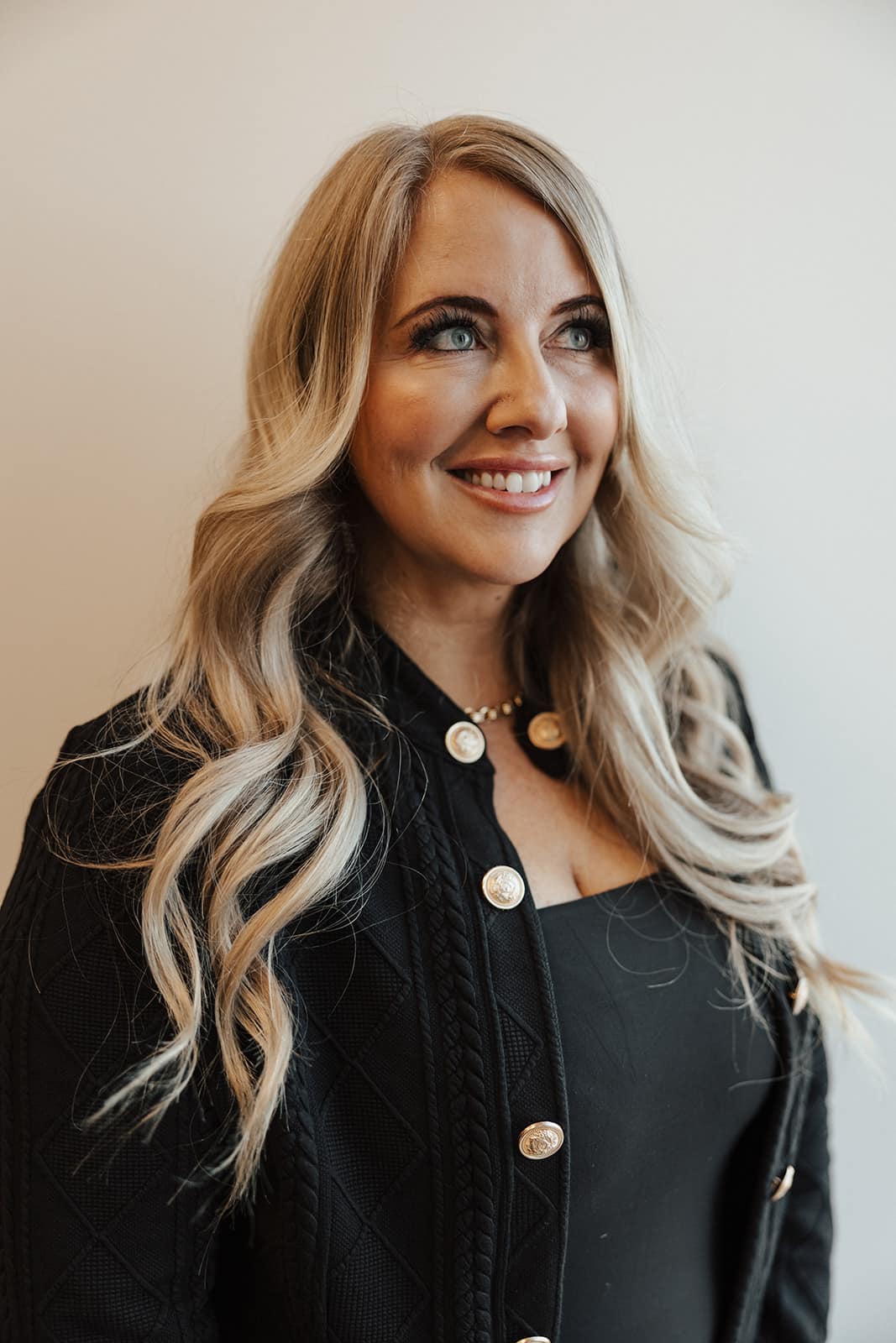 **Alt text:** Portrait of a smiling female patient at Utah Facial Plastics with long blonde hair and radiant, healthy skin. She wears a black outfit with gold-button detailing and looks upward with a confident, refreshed expression, reflecting natural-looking aesthetic results in a warm, modern medical spa setting.