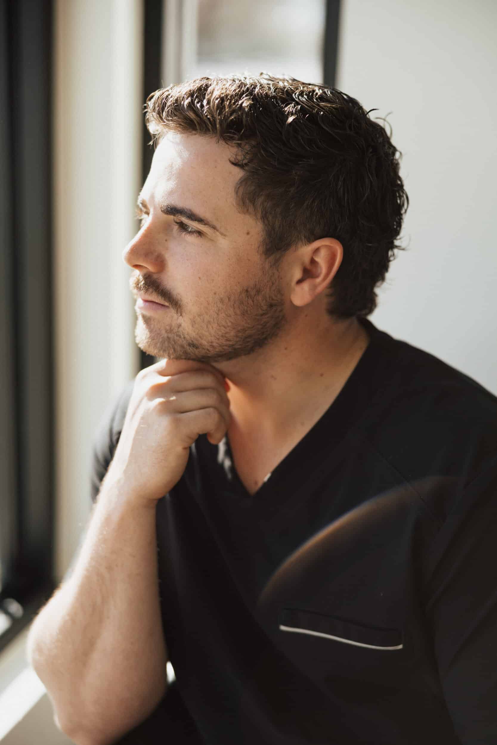 **Alt text:** Portrait of a male patient at Utah Facial Plastics seated by a window in natural light. He rests his chin on his hand with a thoughtful expression, showcasing a strong jawline, healthy skin, and a natural, confident appearance in a modern facial aesthetics practice.
