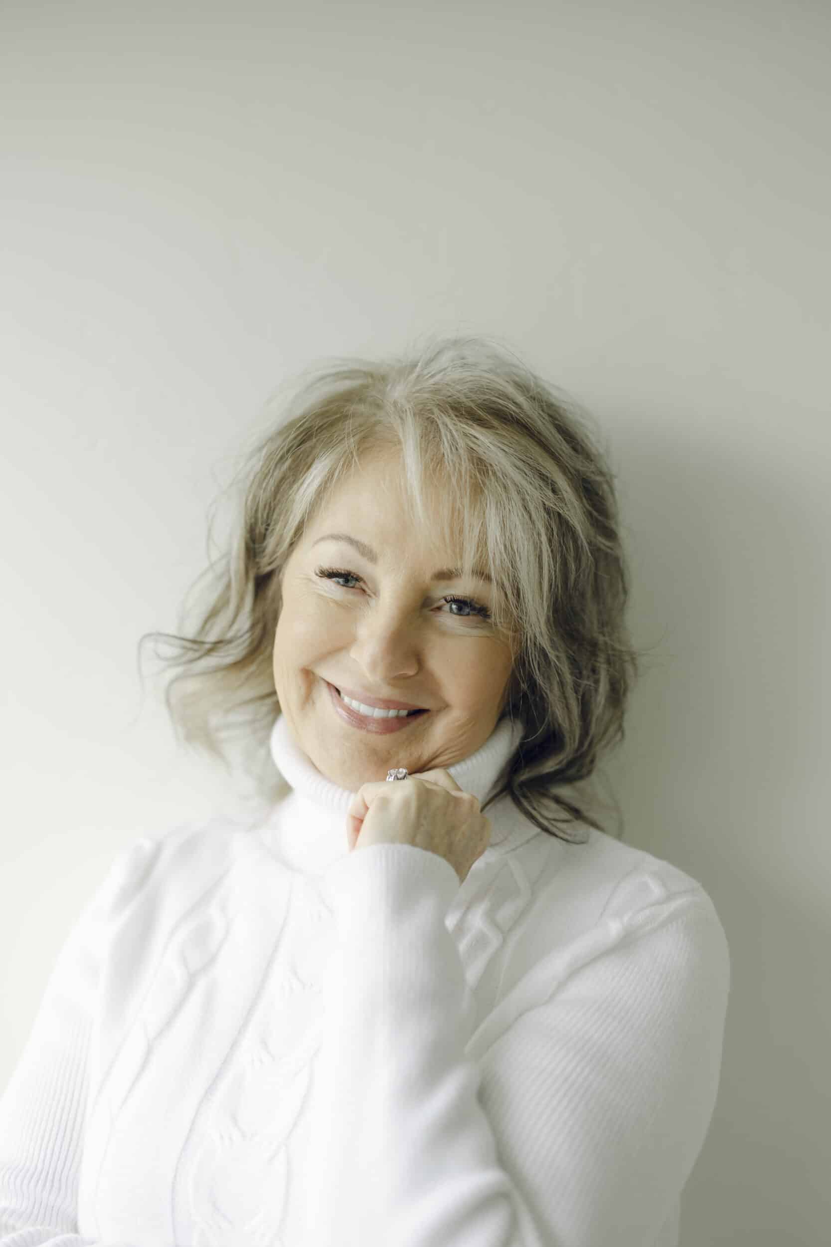 Absolutely. Here's an alt description that aligns with Utah Facial Plastics' warm, elevated, patient-centered brand voice while remaining descriptive and SEO-friendly: **Alt text:** Smiling woman with healthy, radiant skin wearing a white turtleneck sweater. She rests her chin on her hand, showcasing a refreshed, natural appearance that reflects confidence and graceful aging after personalized aesthetic care at Utah Facial Plastics.